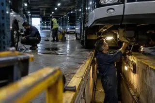 La nuit dans un dépôt de bus : un mécanicien dans la fosse sous un véhicule, un collègue contrôle un pneu