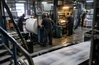De nuit dans une imprimerie, deux conducteurs de rotative installent une bobine de papier, un collègue règle la plieuse