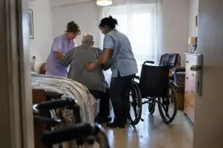 Deux aides à domicile, debout, aident un homme âgé vu de dos à se lever du bord de son lit, un fauteuil roulant vide à côté