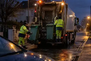 Avant l’aube, deux éboueurs accrochent des bacs à la benne d’un camion de collecte dans une rue de petite ville